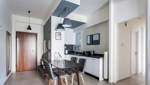 A modern kitchen with white cabinets, a black countertop, and a built-in oven. There is a dining area with a dark wooden table and grey upholstered chairs. - UPSTREET Charming & Comfy 2BD Apt-Acropolis (Athens)
