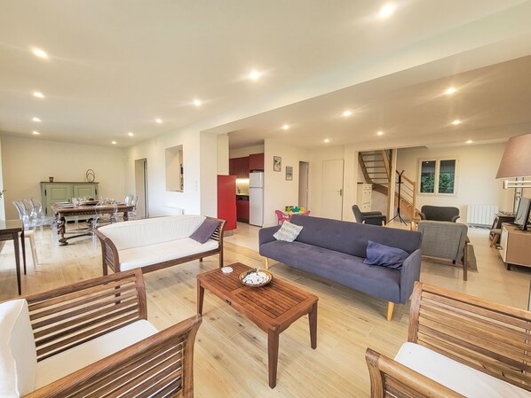 A spacious living area with a sofa, wooden coffee table, and clear chairs. A kitchen is visible in the background with a red refrigerator.