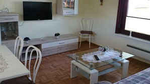 Una sala de estar con televisor, una mesa de centro de cristal, un sofá blanco y un comedor con mesa y sillas.
