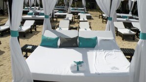 A beachside hotel room with a canopy bed, multiple lounge chairs, and a sandy beach in the background.