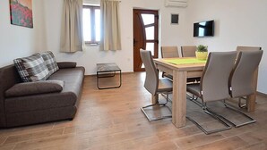 Una habitación amoblada con sofá, una mesa de comedor con sillas, una mesita, un televisor y un ventanal con cortinas.