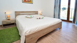 Un dormitorio con una cama de madera, dos mesitas de noche con lámparas, un tapete verde, un cuadro en la pared y una puerta corrediza de vidrio al exterior.