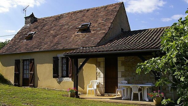 Ein rustikales Haus mit Ziegeldach, einer kleinen Veranda mit Gartenmöbeln und einem grünen Rasen.