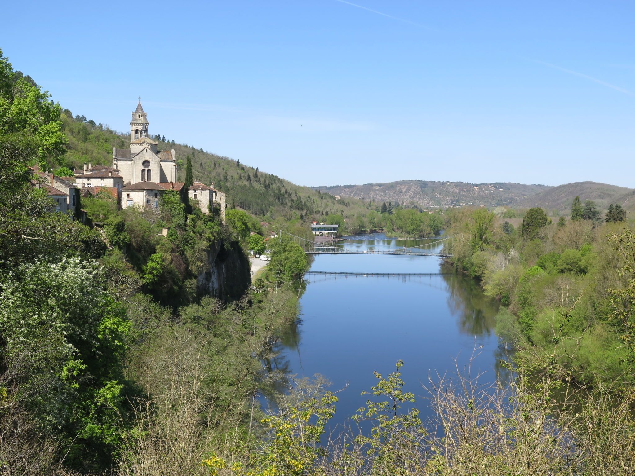 Une église historique dotée d’un clocher, entourée d’une végétation luxuriante et bordée d’une rivière paisible.