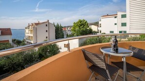 Un balcón con mesa y sillas, con vista a una zona costera con edificios y árboles.