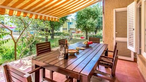 An outdoor patio with a wooden table and chairs, a striped awning, and a view of a garden and buildings in the background. - Apartment Silva by Interhome (Fažana/Peroj)