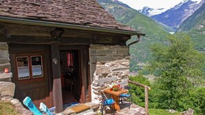 Un rustico edificio in pietra con una porta di legno, un piccolo portico con tavolo e sedie, e vista su una valle con montagne sullo sfondo.