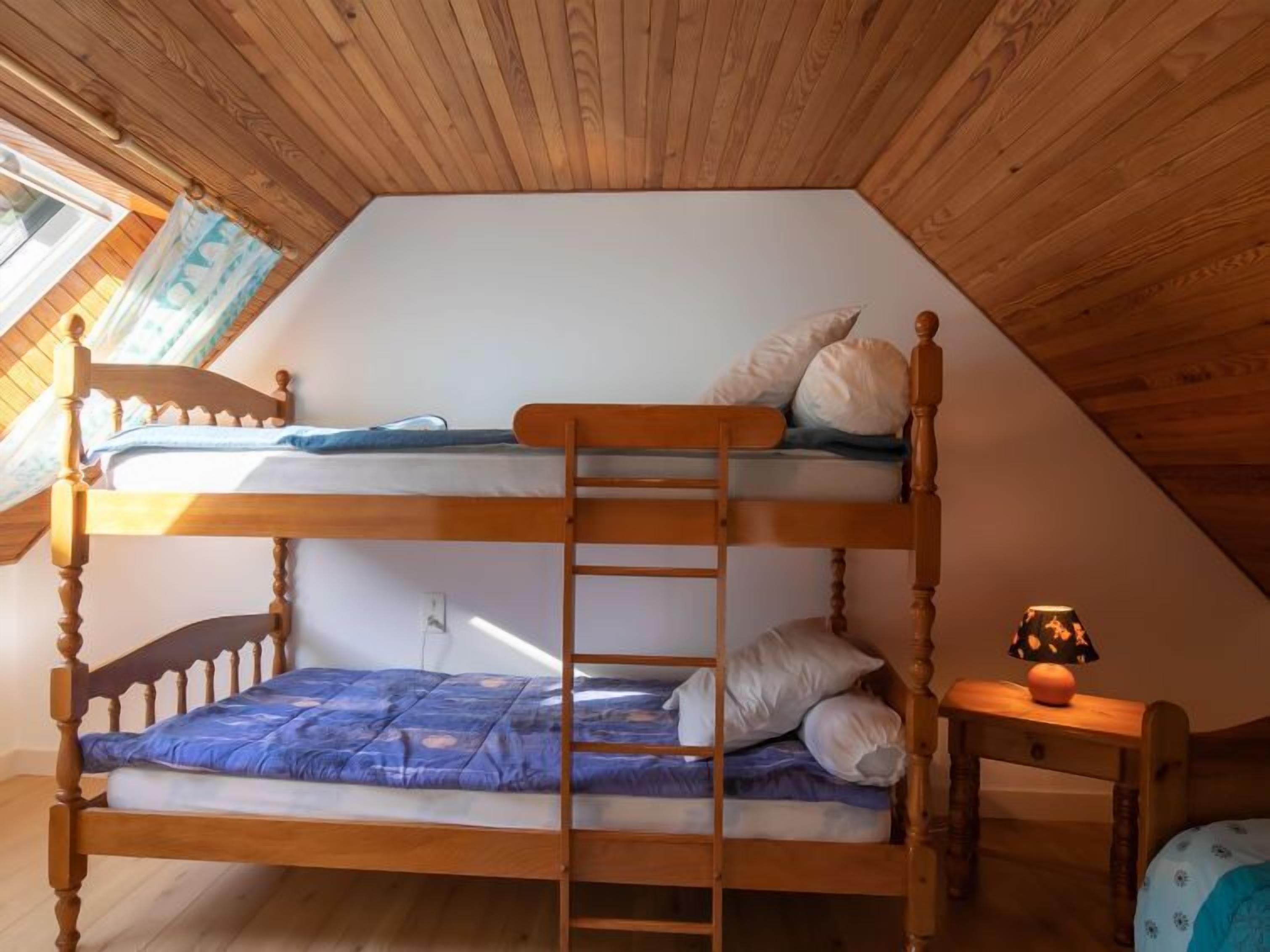 A dormitory-style room with two bunk beds, wooden ceiling beams, a window with curtains, and a bedside table with a lamp.
