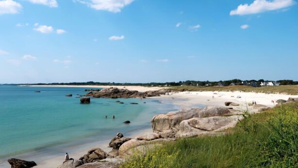 A coastal beach with clear blue water, large rocks, and a distant shoreline with buildings. - Ty Pendruc by Interhome (Tregunc)