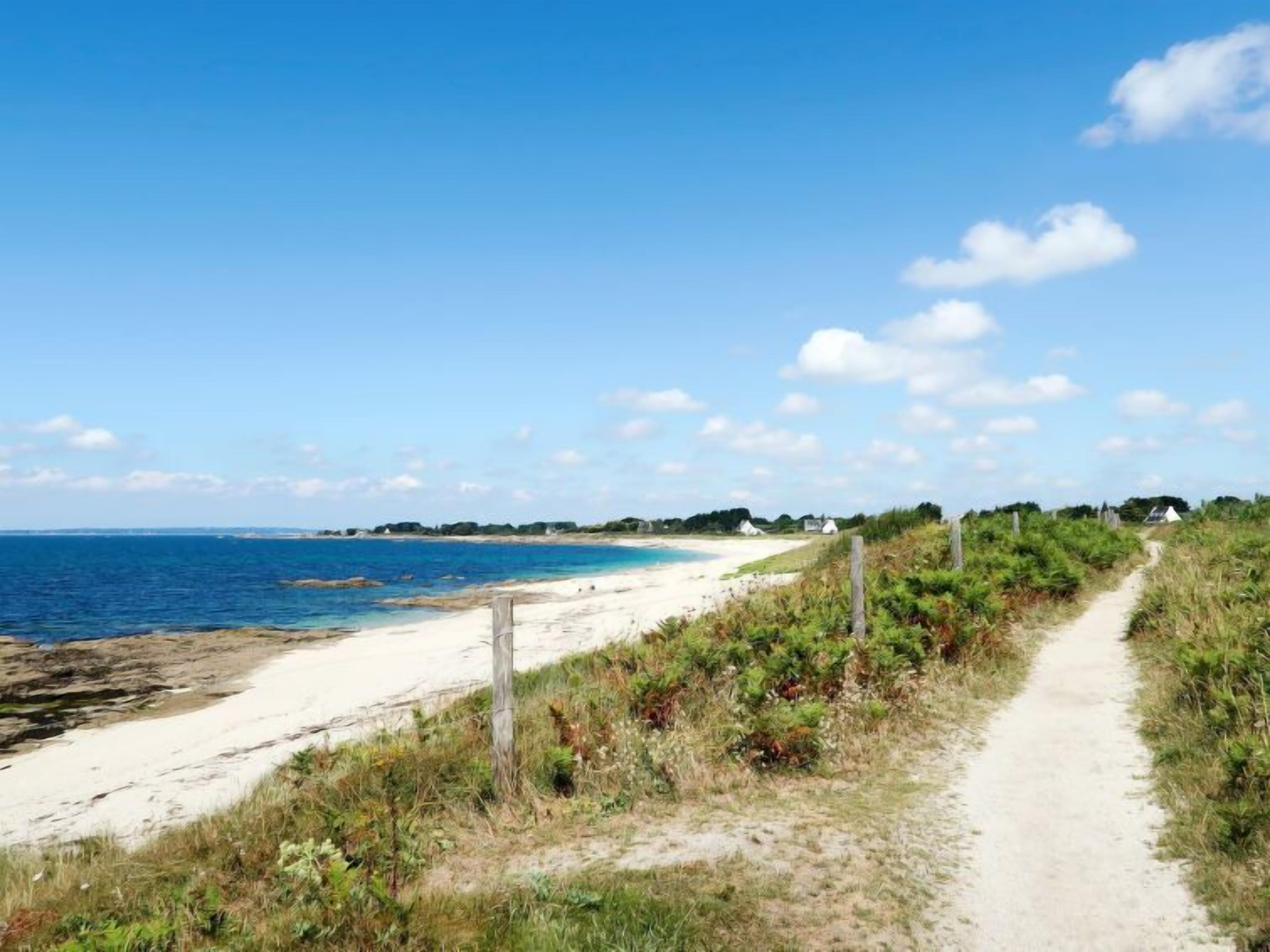 Un sentier côtier mène à une plage où l’eau est d’un bleu limpide, et où l’on aperçoit quelques maisons au loin.