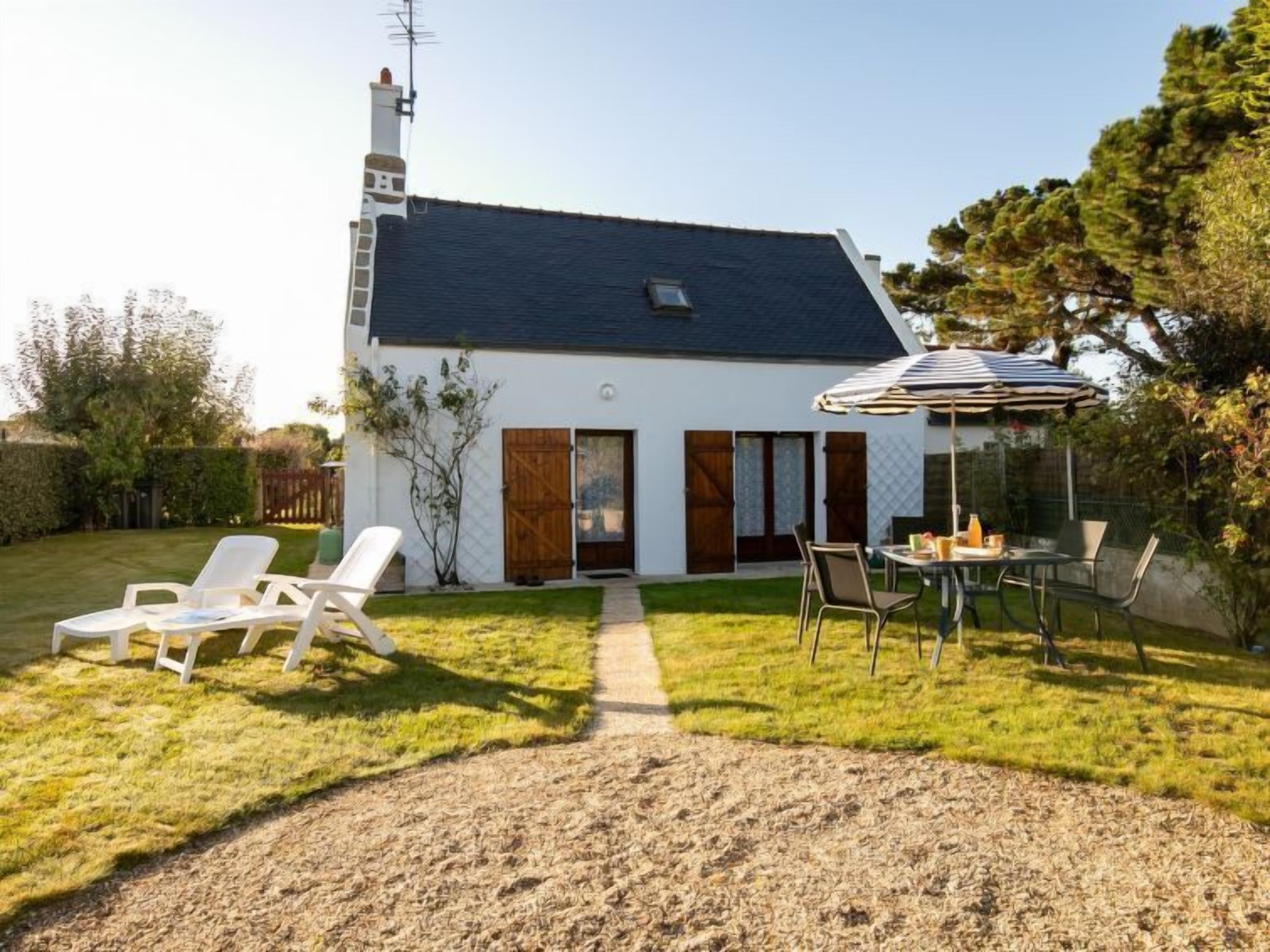 Una casa con techo oscuro, paredes exteriores blancas y un pequeño porche delantero. Cuenta con un patio con mesa y sillas, además de un espacio de estar al aire libre cubierto.