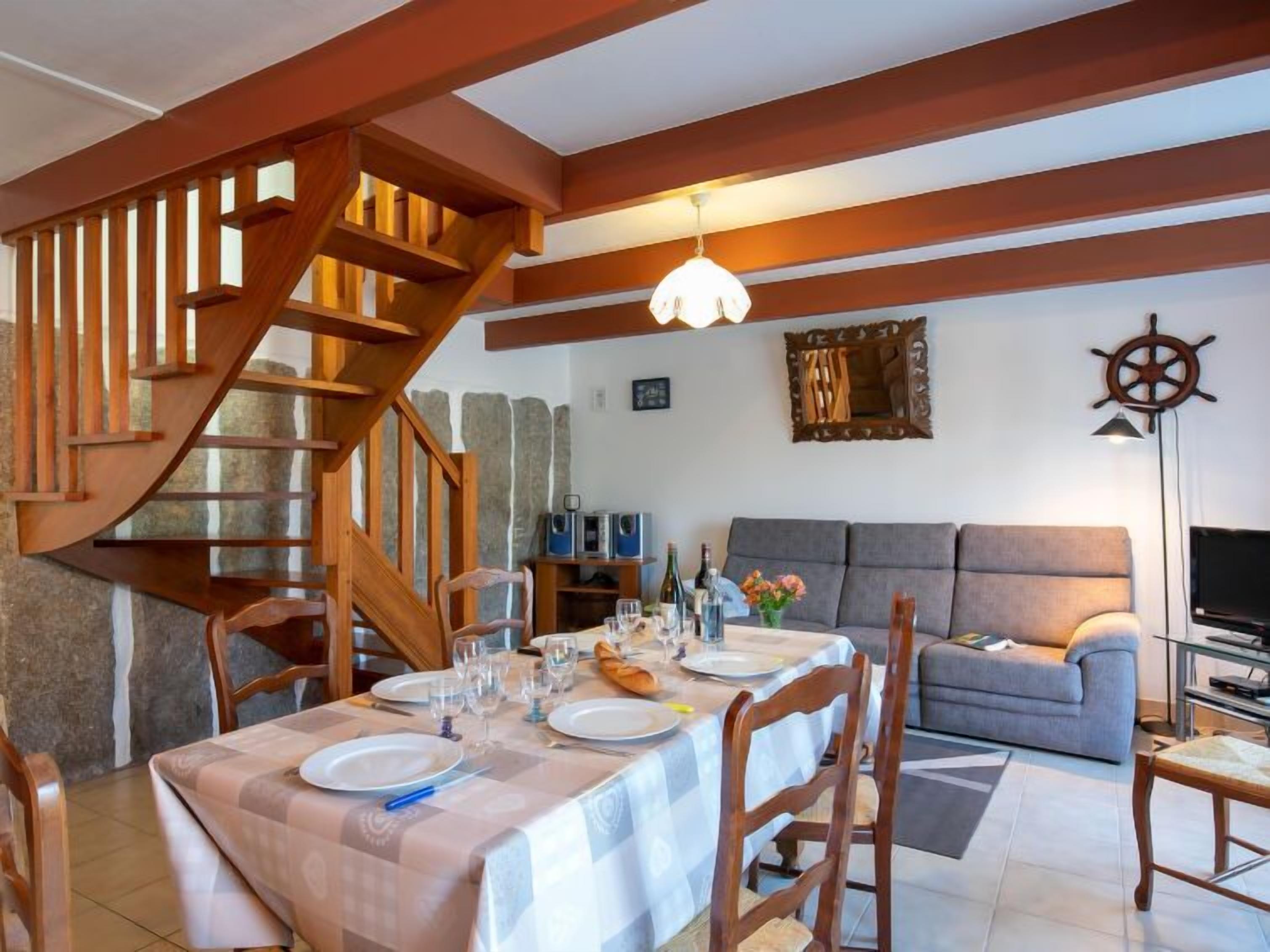A dining area with a table set for four, a wooden staircase, a grey sofa, and a television in the living area.