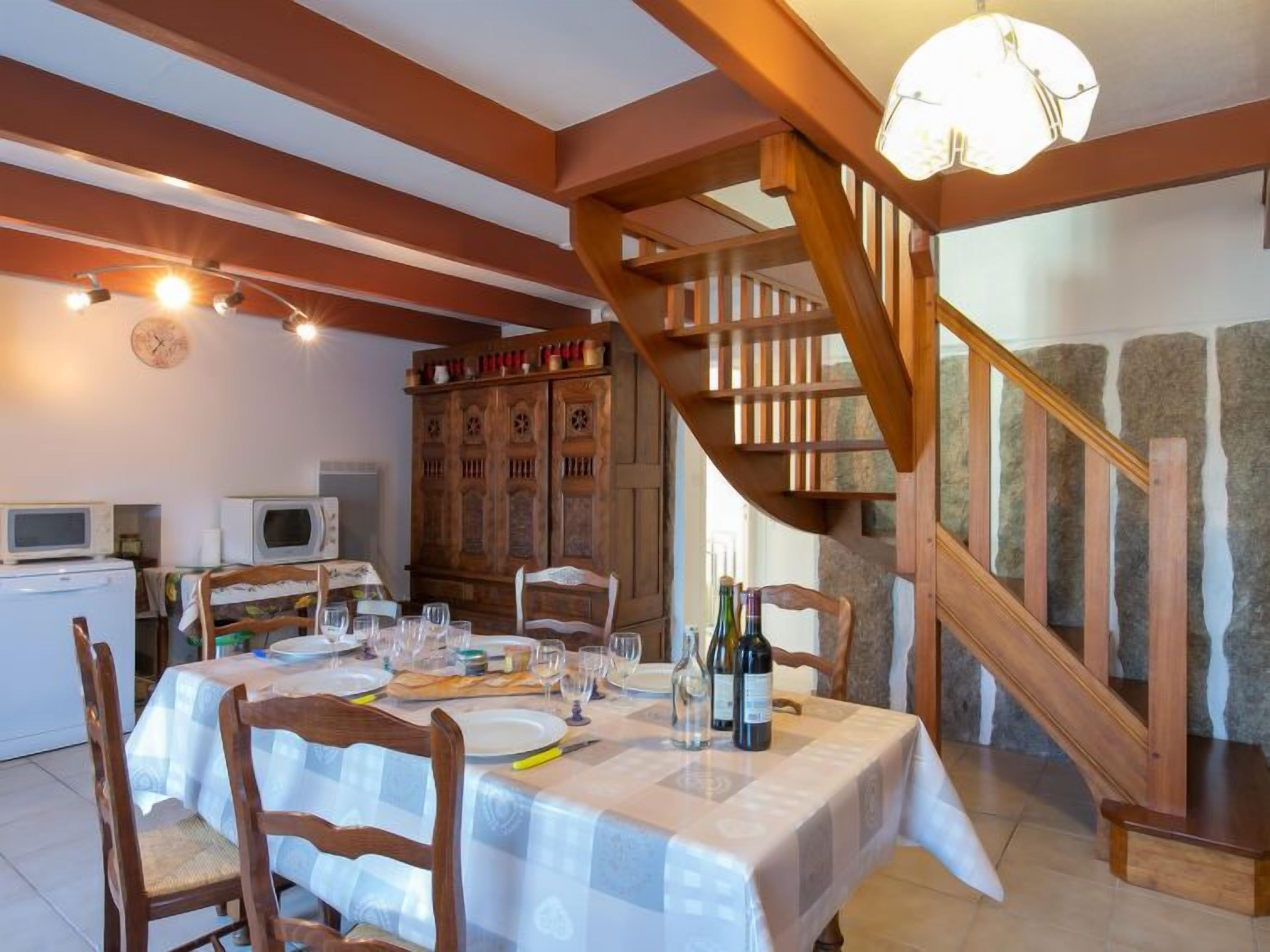Un coin repas avec une table dressée pour quatre personnes, des chaises en bois, un escalier en bois et un mur en pierre.