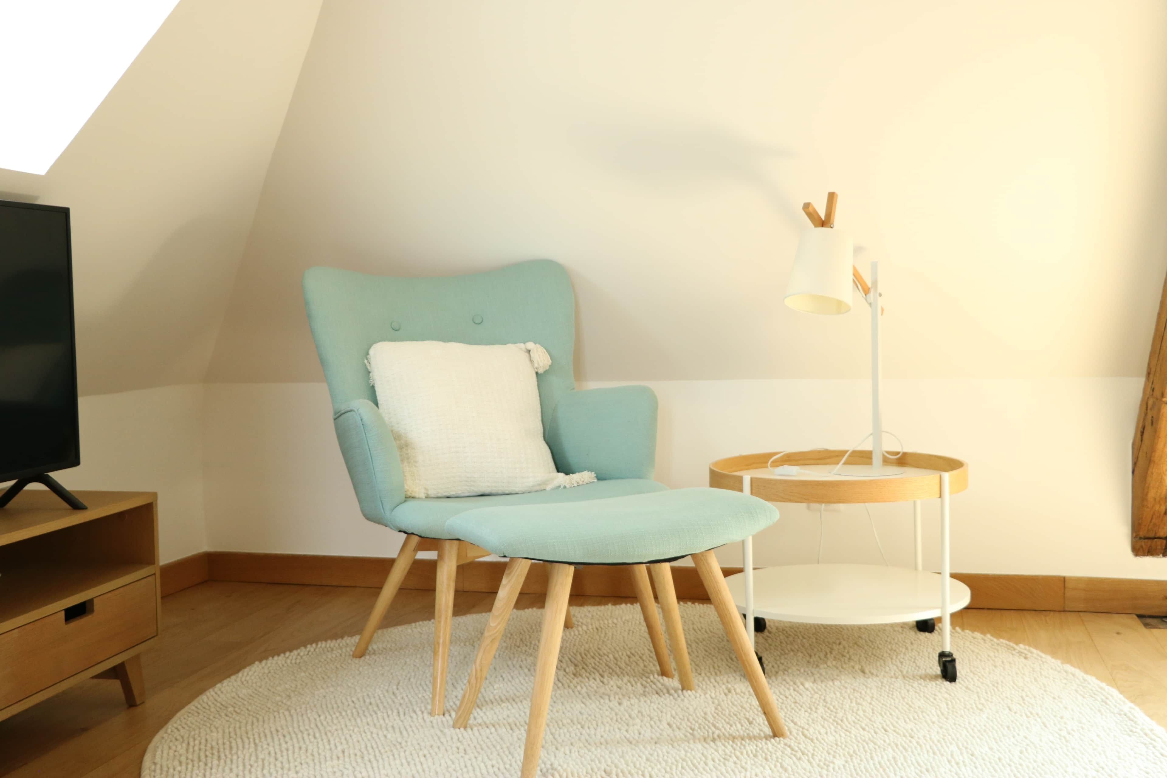 A room with a teal armchair, a white throw pillow, a small side table with a lamp, and a television on a wooden stand.