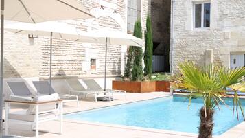 A poolside area with lounge chairs and umbrellas, a stone building in the background, and a palm tree.