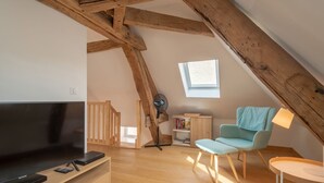 Ein gemütliches Dachzimmer mit Holzbalkendecke, einem großen Fernseher, einem blauen Sessel und einem kleinen Tisch.