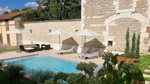 Ein Poolbereich mit Liegestühlen und Sonnenschirmen, im Hintergrund ein Steinhaus und im Vordergrund üppiges Grün.