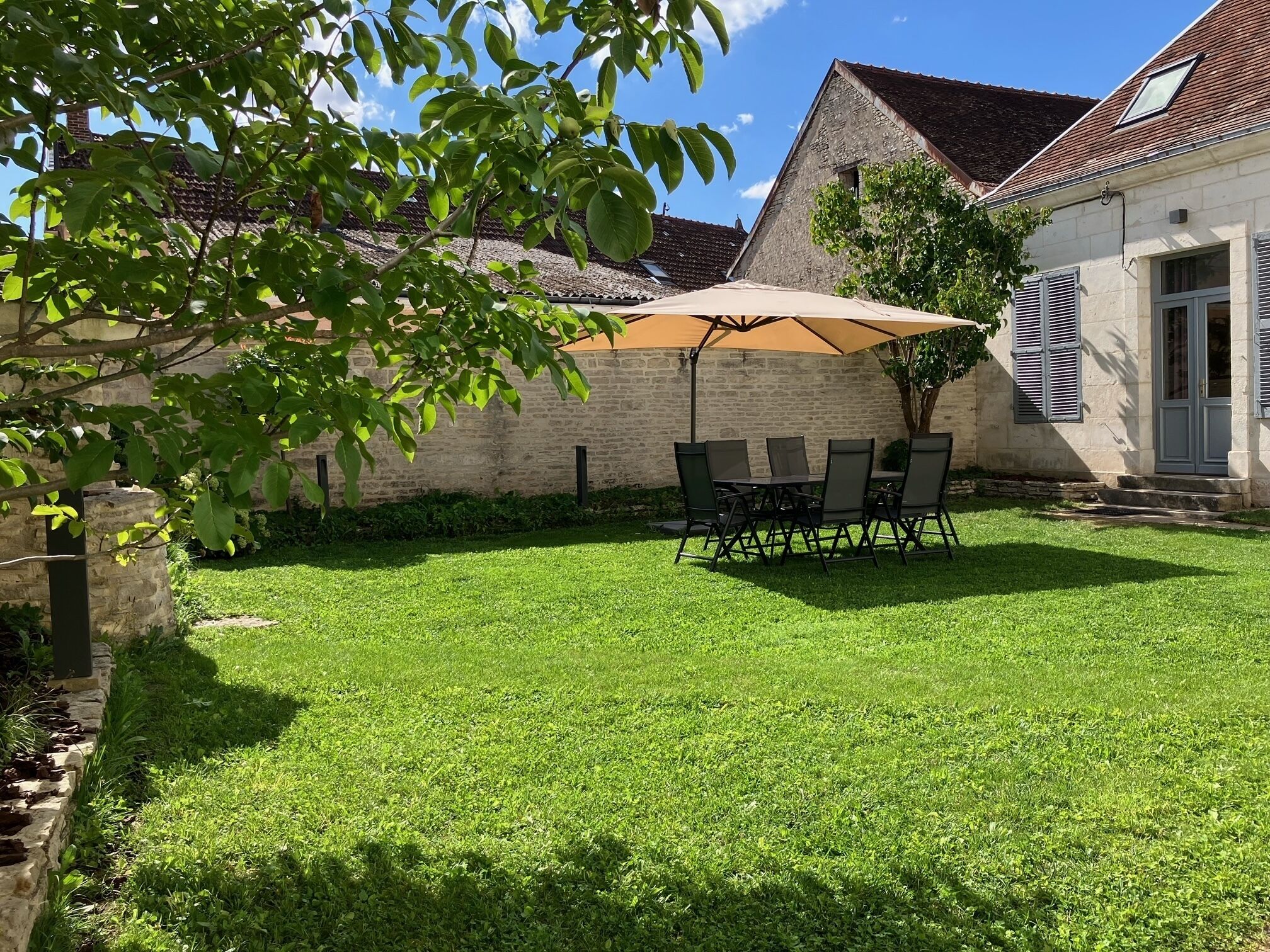 Une terrasse bien entretenue, avec une table et des chaises placées sous un grand parasol, entourée d’un mur de pierres et d’une pelouse verdoyante.