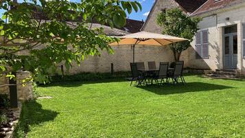 Une terrasse bien entretenue, avec une table et des chaises placées sous un grand parasol, entourée d’un mur de pierres et d’une pelouse verdoyante.