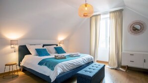 Ein Schlafzimmer mit Bett, Nachttischlampen, einer Hängeleuchte, einem Fenster mit Vorhängen, Holzboden und einem wandmontierten Spiegel.