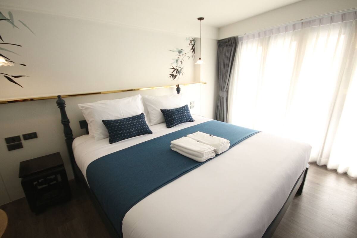 Una cama bien hecha con colcha azul y almohadas blancas, una mesita de noche pequeña y una lámpara de pared.