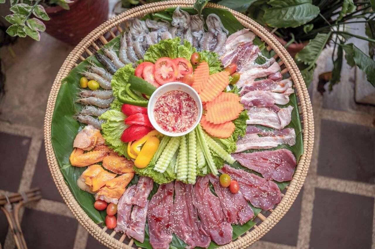 Una cesta grande y redonda, tejida, llena de diversos mariscos y verduras, incluyendo camarones, pescado y vegetales en rodajas, dispuesta sobre una hoja verde.