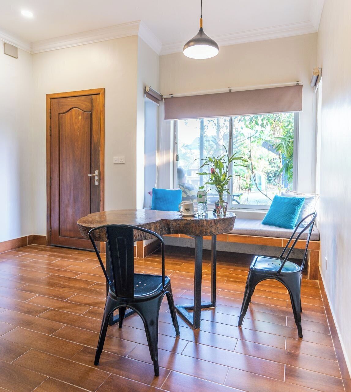 Comedor con una mesa redonda de madera, dos sillas negras, una planta en maceta y una ventana con vistas al verdor.