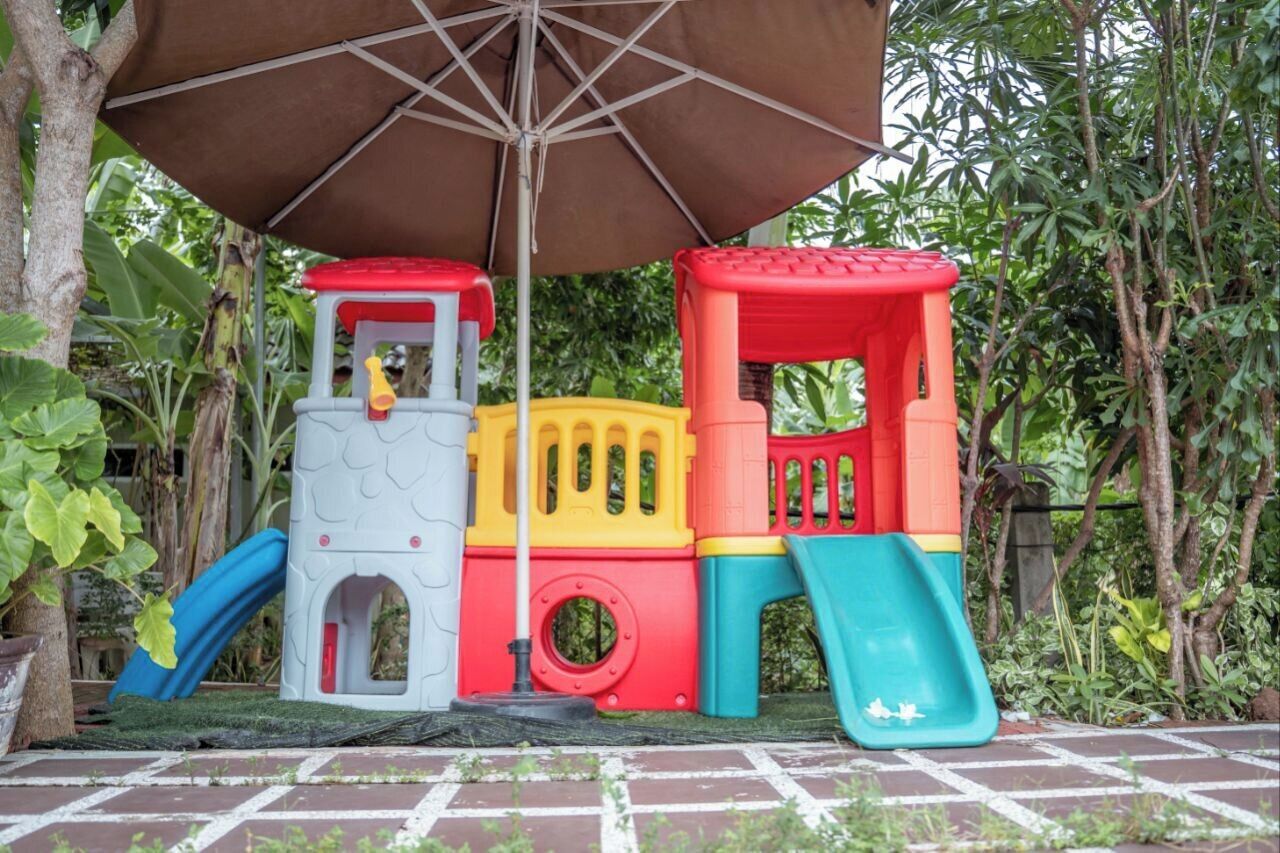 A children's play structure with slides and climbing elements, set on a brick patio surrounded by greenery.