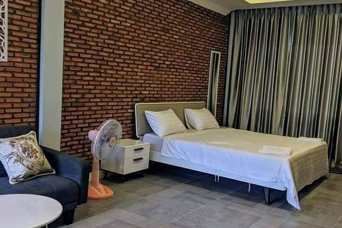 A bedroom with a brick wall, a bed, a nightstand, a fan, a sofa, and a small table.