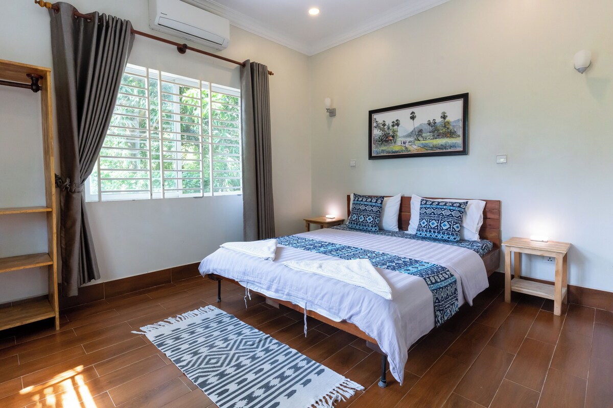 A bedroom with a bed, a nightstand, a wooden shelf, a window with curtains, and a painting on the wall.