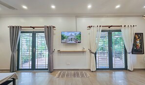 A room with two large sliding glass doors, a flat-screen TV mounted on the wall, a small shelf with a decorative item, and a patterned rug on the floor. - Sanssouci Kep (Kep)