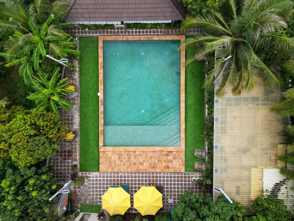An aerial view of a rectangular swimming pool with a wooden deck surrounding it, bordered by green grass and various tropical plants. - Sanssouci Kep (Kep)