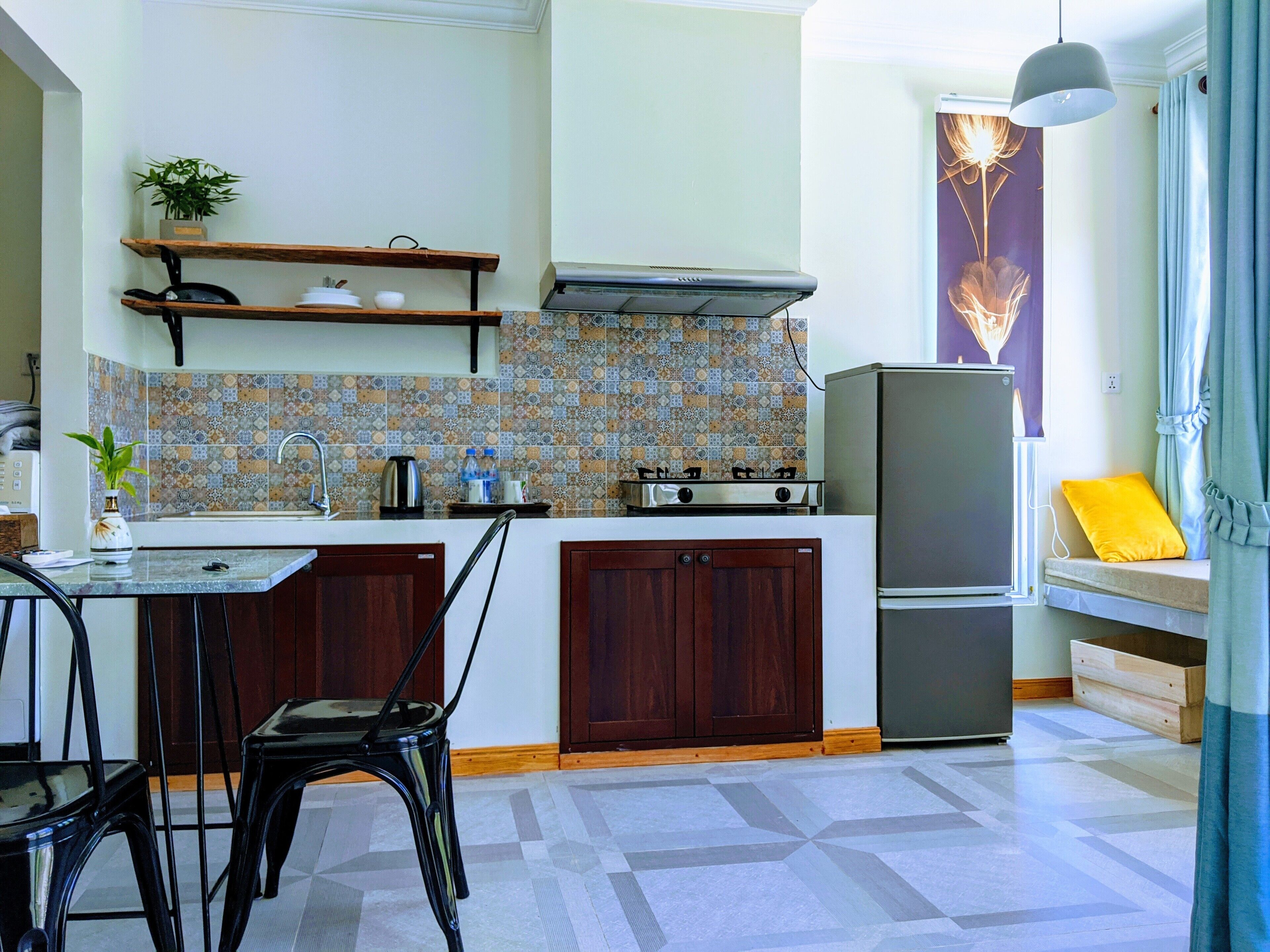 A compact kitchen with a gas stove, a sink, and a refrigerator. There is a dining table with chairs and a shelf above the counter.