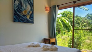 A bedroom with a large bed, a small table, a painting on the wall, and a view of greenery through a window. - Sanssouci Kep (Kep)