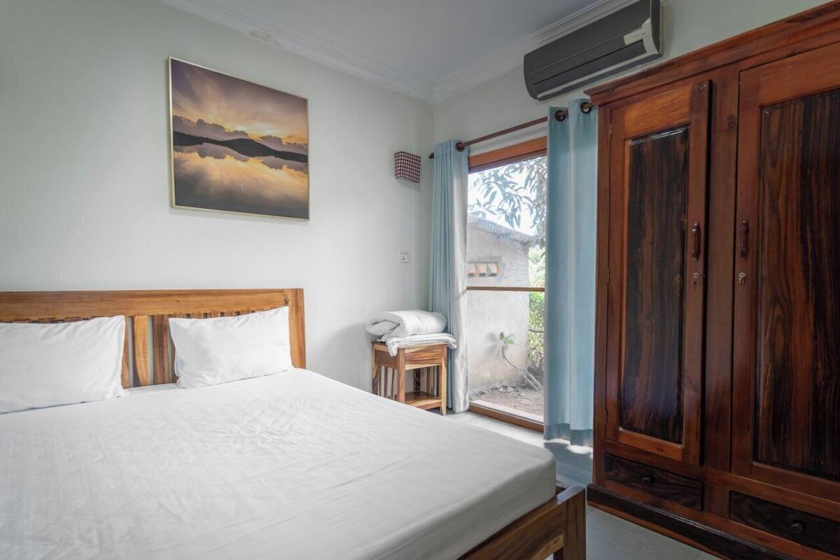 A bedroom with a large bed, a wooden wardrobe, a small table with a chair, and a window with curtains.