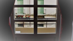A view through a window with a balcony, featuring a building with green shutters and a metal railing. - Domus Claudiae's studio C-1 (Catania)