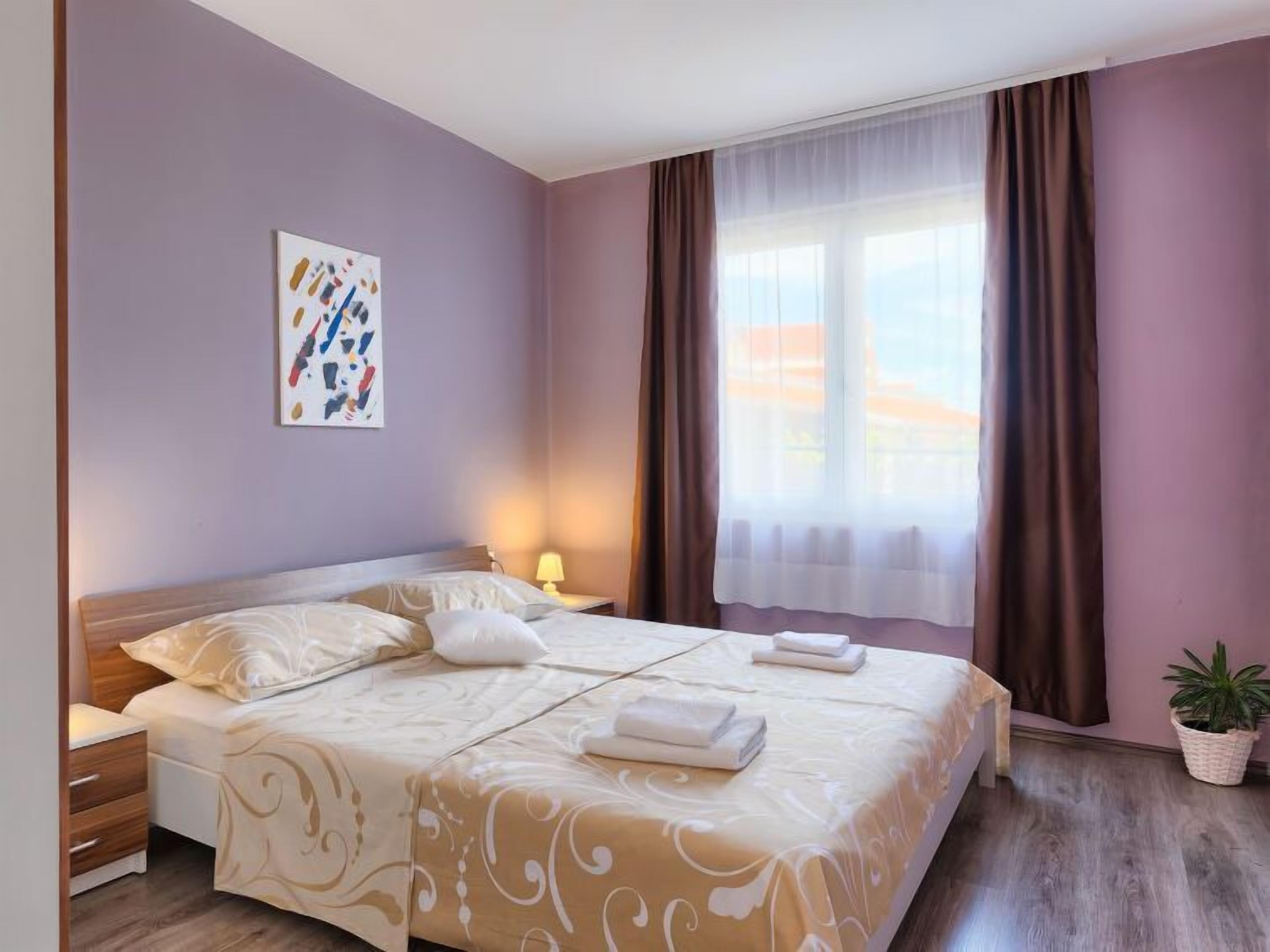 A hotel room with a large bed, a bedside table with a lamp, a window with curtains, and a wall-mounted artwork.