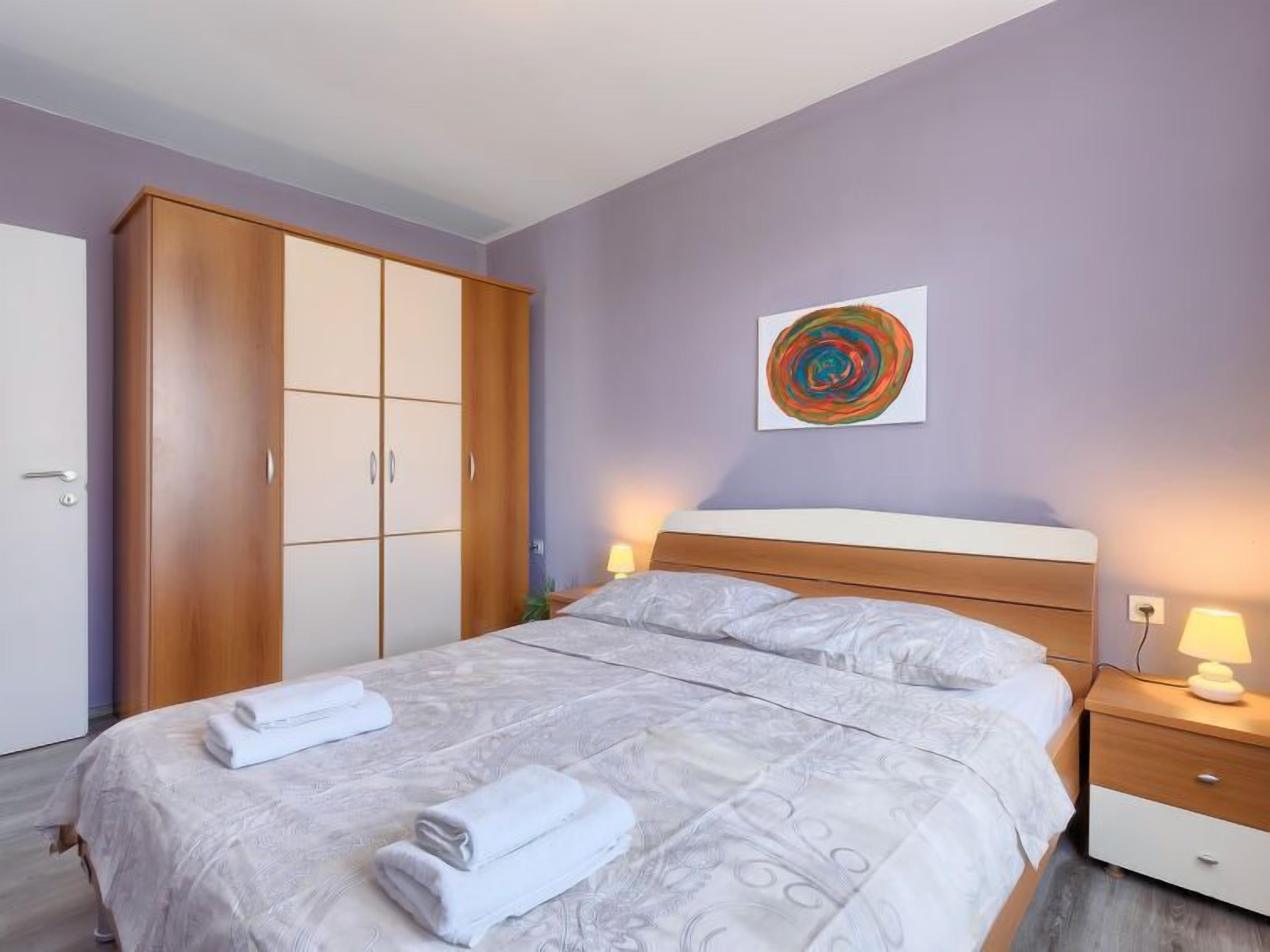 A bedroom with a bed, a wardrobe, a bedside table with a lamp, and a wall art piece.