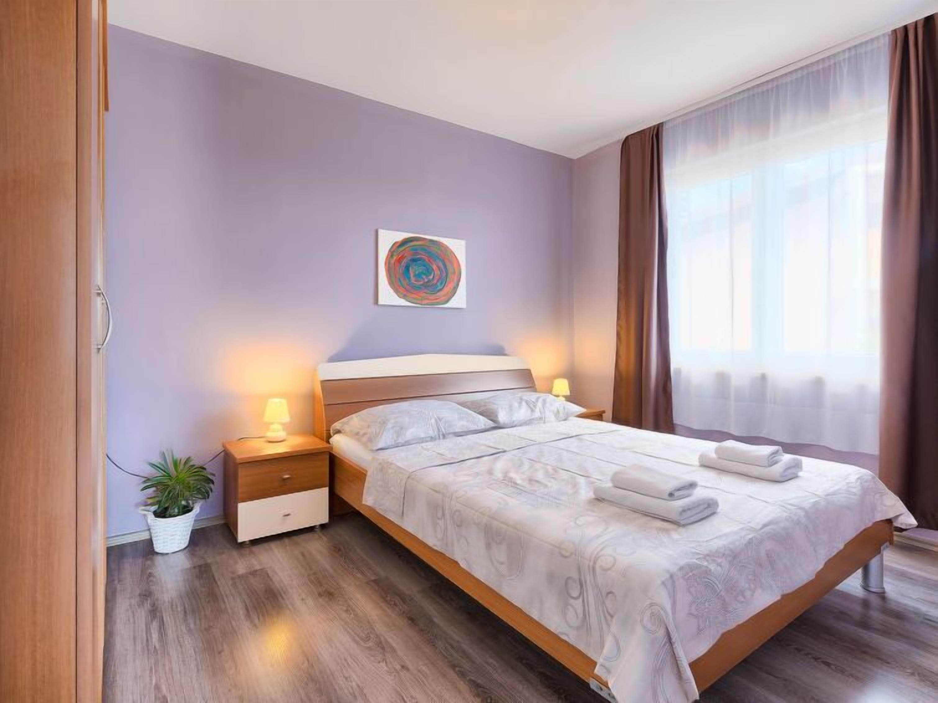 Un dormitorio con cama, mesitas de noche, una planta en maceta y un cuadro en la pared.