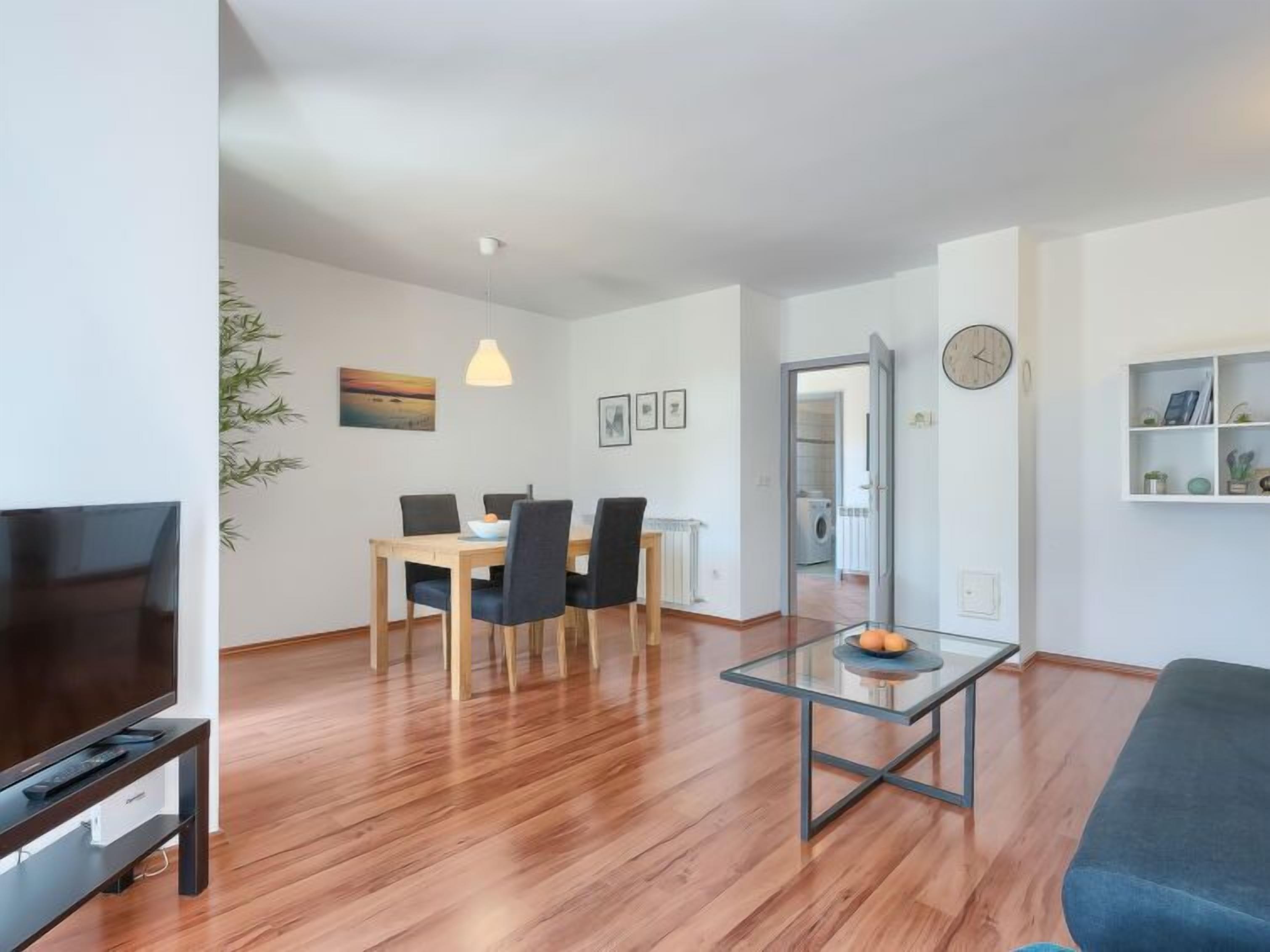 A modern living room with a flat-screen TV, a glass coffee table, and a blue sofa. There is a dining area with a wooden table and chairs, and a kitchen visible through an open door.