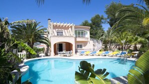 A poolside view of a two-story building with a balcony, surrounded by palm trees and a clear blue sky. - Vacation home Amber by Interhome (Calpe/Calp)