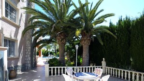 An outdoor patio with a round table and four chairs, surrounded by a white balustrade. Palm trees and a hedge are in the background. - Vacation home Amber by Interhome (Calpe/Calp)