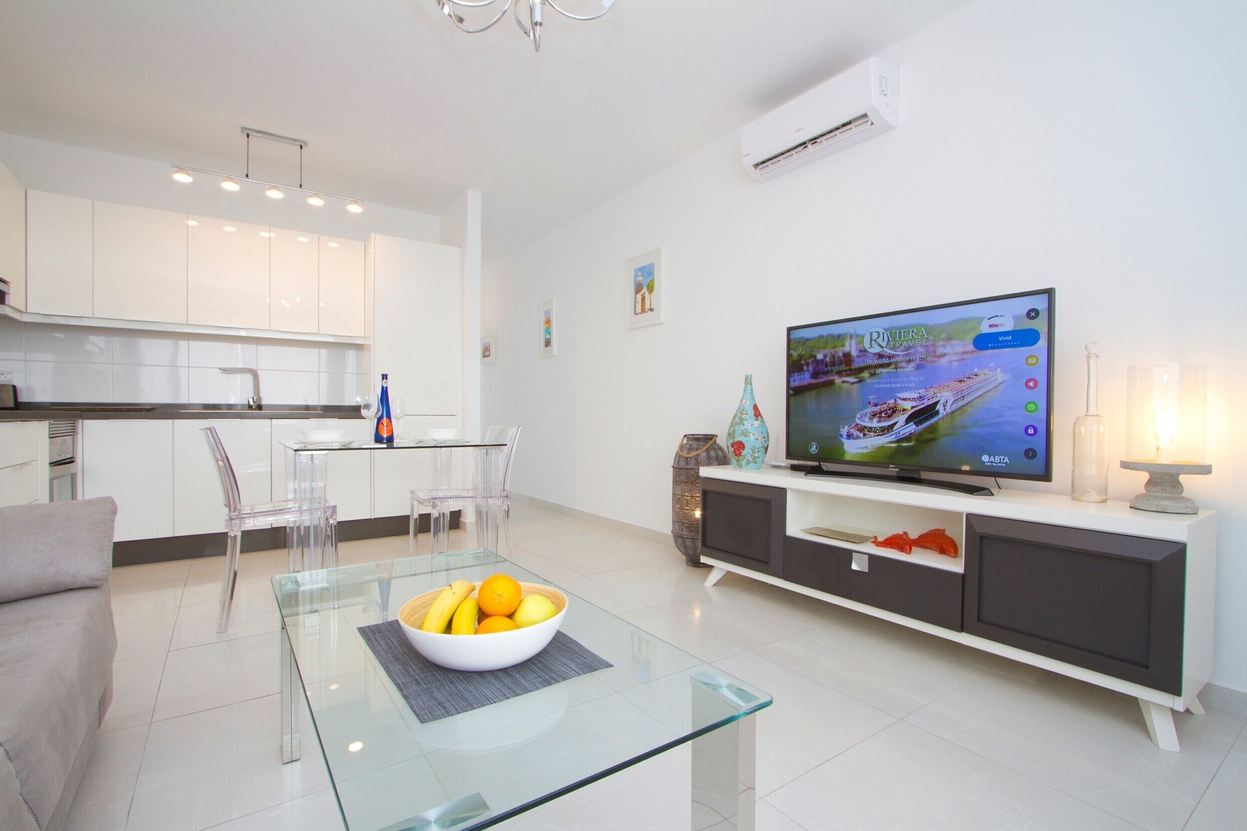 A modern living room with a glass coffee table, a flat-screen TV, and a dining area with a glass table and chairs.