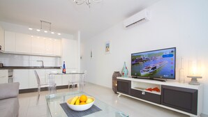 Una sala moderna con una mesa de centro de vidrio, un televisor y una zona de comedor con un armario blanco y sillas transparentes.