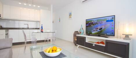 A modern living room with a glass coffee table, a flat-screen TV, and a dining area with a glass table and chairs.