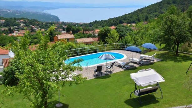 A swimming pool with lounge chairs and umbrellas, surrounded by a well-maintained lawn and trees, with a view of a lake and distant hills. - Vacation home Lea by Interhome (Opatija/Rukavac)