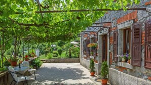 A covered outdoor patio with a table and chairs, surrounded by potted plants and a stone building with wooden shutters. - Vacation home Lea by Interhome (Opatija/Rukavac)