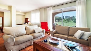 Een ruime woonkamer met twee banken, een rode fauteuil en een glazen salontafel. Een groot raam biedt uitzicht op een groen landschap.