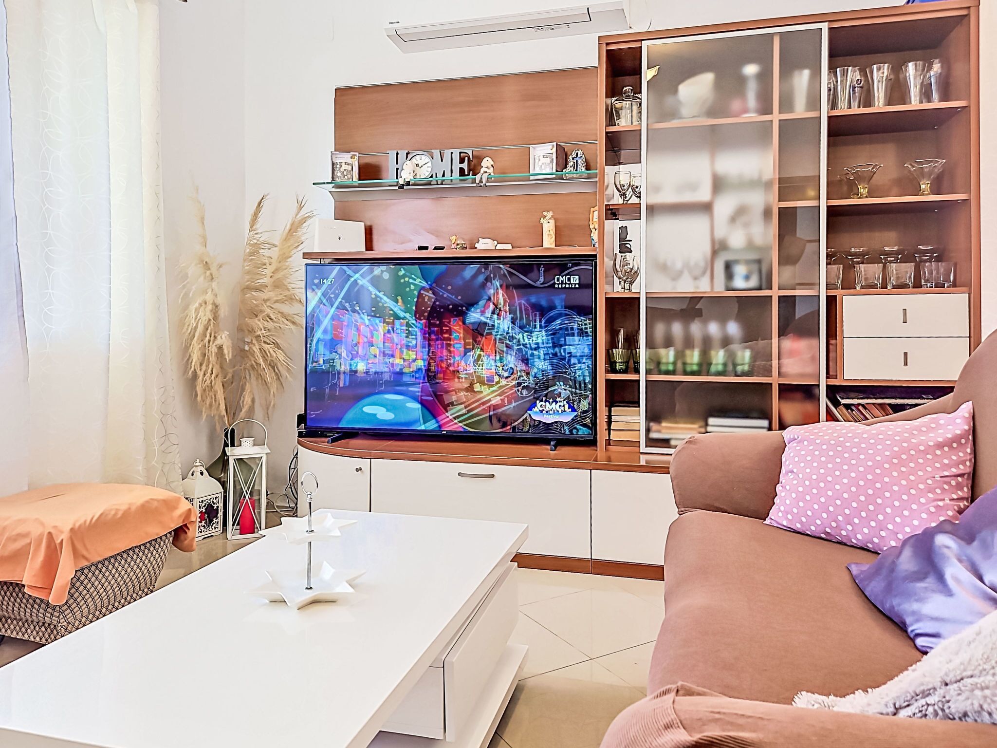 Ett vardagsrum med en platt-TV, ett vitt soffbord, en soffa med kuddar i prickigt mönster och en bokhylla i trä med dekorativa föremål.