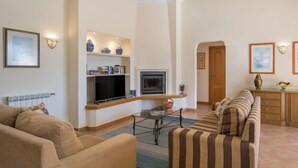 Een woonkamer met een bank, een televisie en een open haard. Er staan planken met decoratieve objecten en een deur die naar een andere kamer leidt.